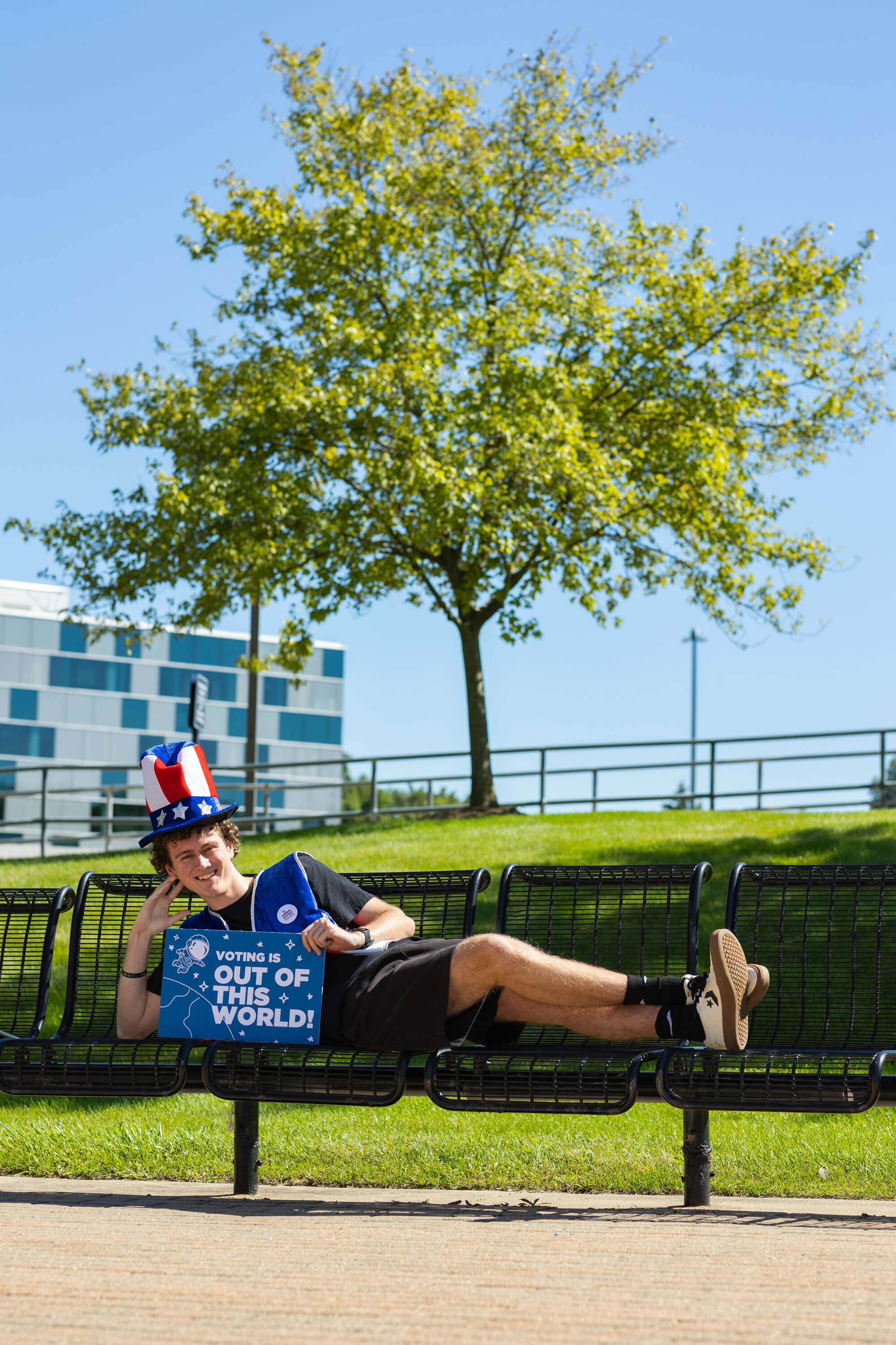 A student wearing an Uncle Sam hat, sitting on a bench, and holding a sign that says "voting is out of this world!"